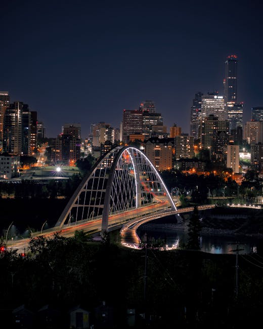 A nighttime aerial view of a city skyline with numerous illuminated high-rise buildings, and a modern, illuminated arch bridge spanning a river. The scene shows the bridge's curved design with metal f