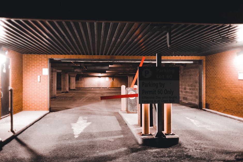 A narrow residential street at the entrance to a multi-storey apartment complex, featuring parked cars on both sides including white, black, and dark grey vehicles. The road surface appears wet, and a